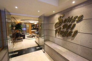 Zona de recepción de un hotel con un separador de vidrio, mostrador de recepción y una pared con el nombre del hotel.