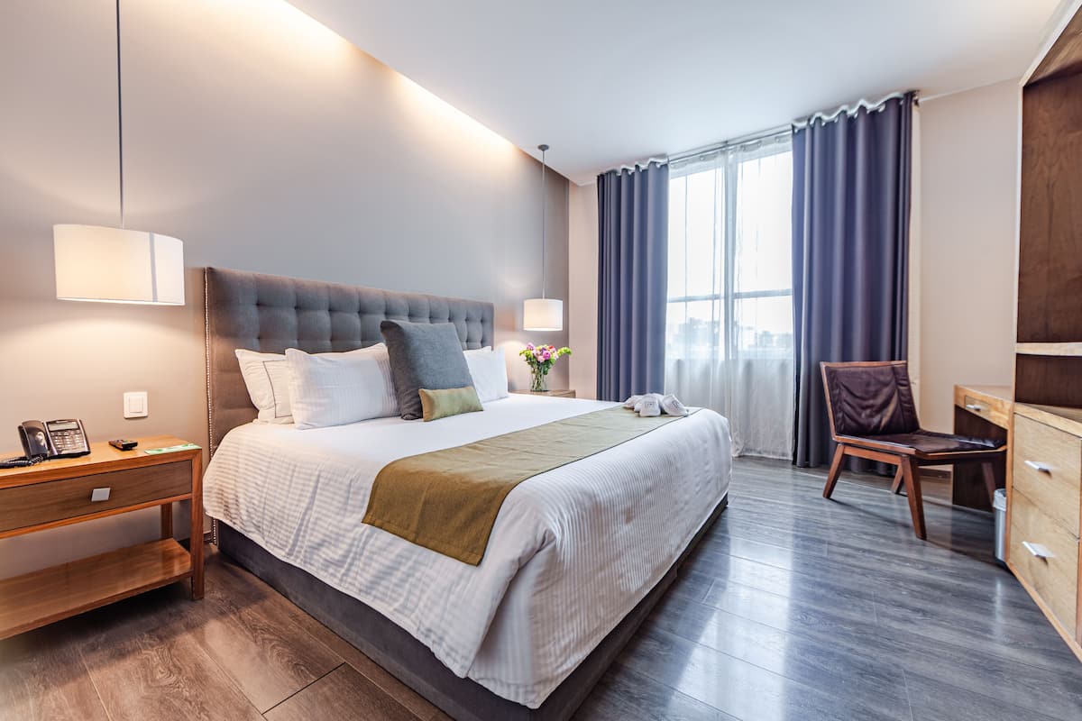 A modern hotel room with a large bed, bedside table, lamp, and a view of the city through the window.