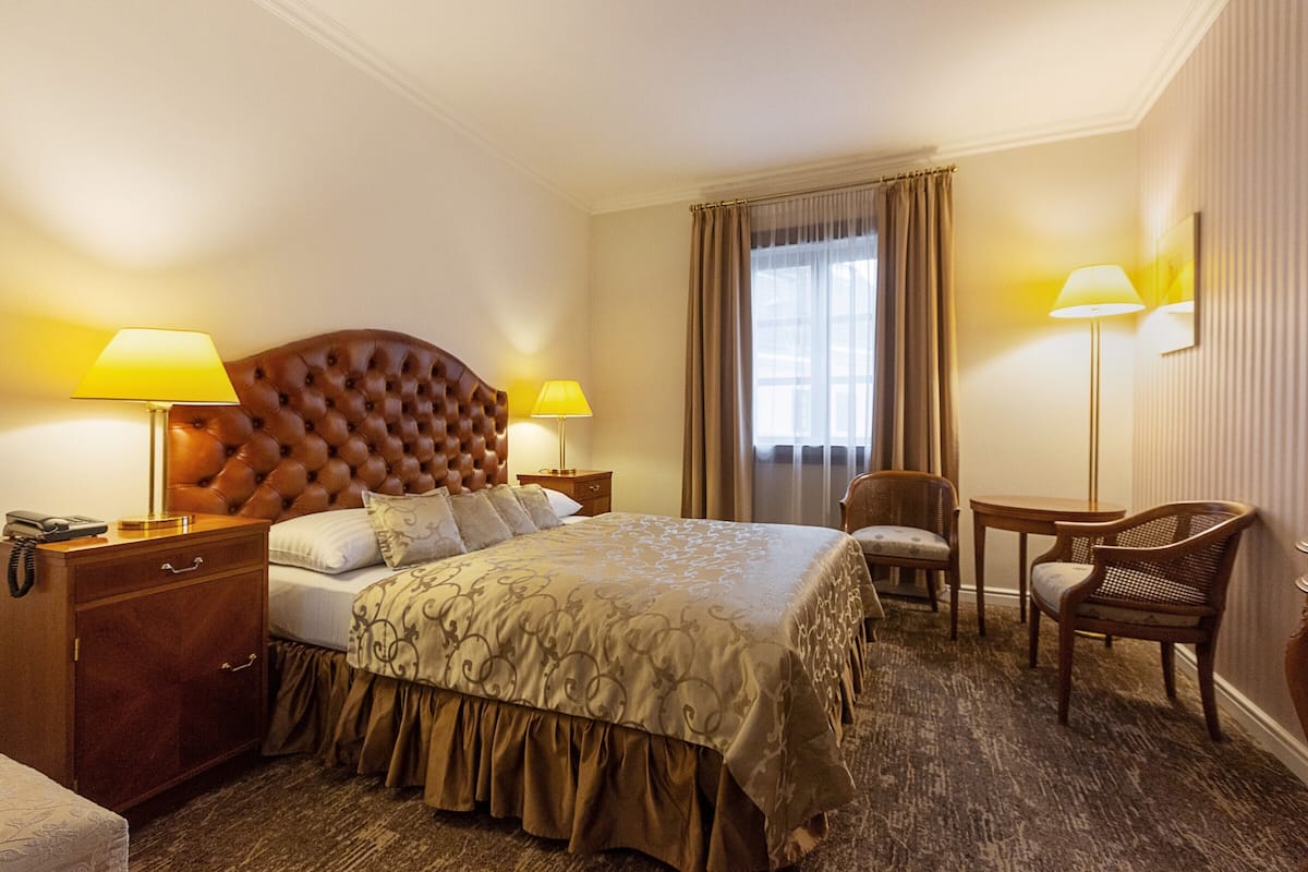 A hotel room with a tufted headboard bed, two bedside lamps, a wooden nightstand, a desk with a chair, and a large window with curtains.