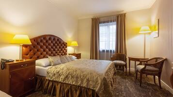Ein Hotelzimmer mit einem bettbezogenen Kopfteil, zwei Nachttischlampen, einem Holznachttisch, einem Schreibtisch mit Stuhl und einem großen Fenster mit Vorhängen.