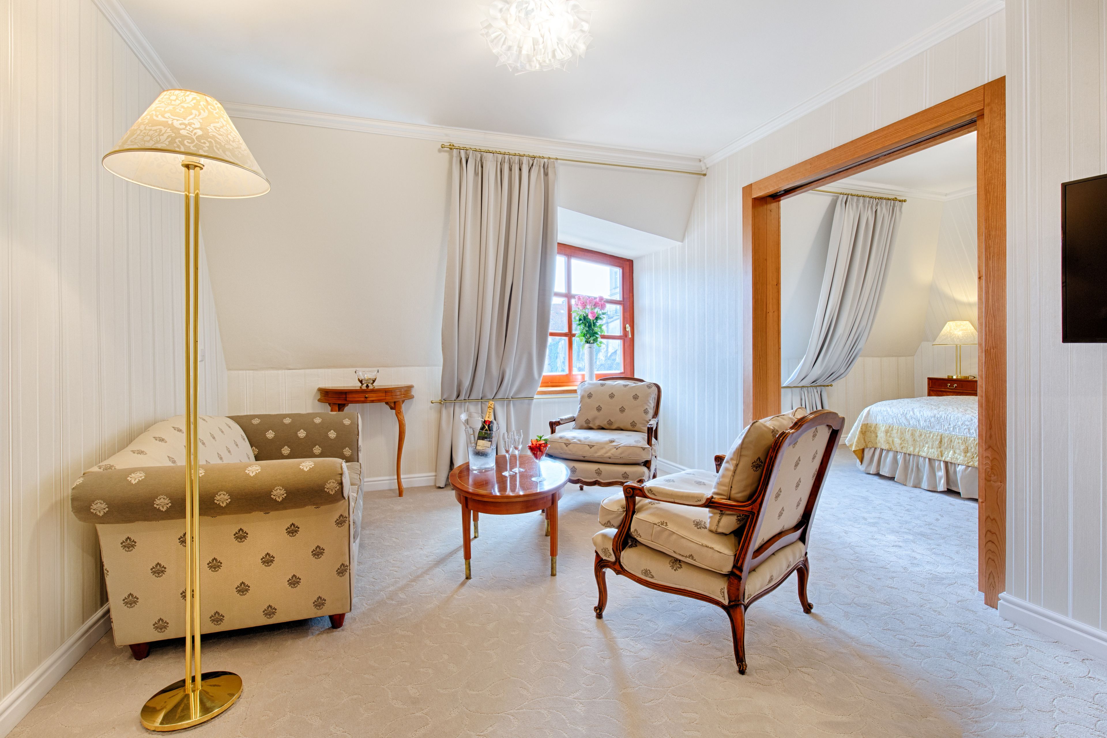 Ein Hotelzimmer mit einer Couch, einem Sessel und einem kleinen Tisch mit Lampe. Im Hintergrund ist ein Bett zu sehen und ein Fenster mit Vorhängen.