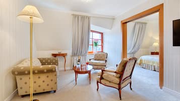Ein Hotelzimmer mit einer Couch, einem Sessel und einem kleinen Tisch mit Lampe. Im Hintergrund ist ein Bett zu sehen und ein Fenster mit Vorhängen.