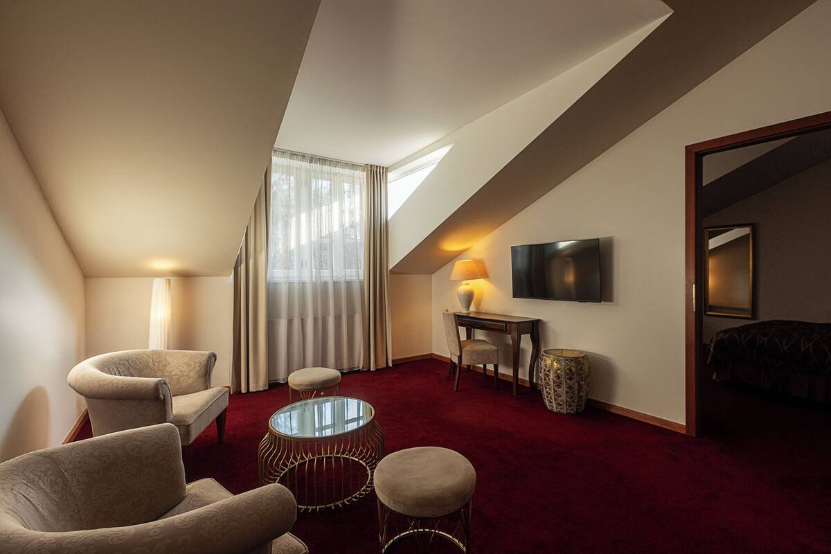 A hotel room with a red carpet, two armchairs, a glass coffee table, a desk with a lamp, a TV on the wall, and a bed.