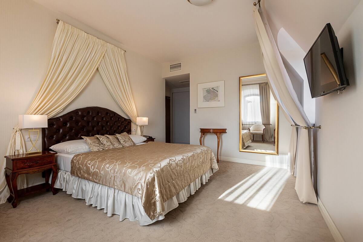 A bedroom with a large bed, a bedside table with a lamp, a mirror, and a television mounted on the wall.