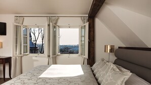 A bedroom with a large bed, a nightstand, a lamp, and a view of the city through open windows. - Hotel Hoffmeister (Prague)