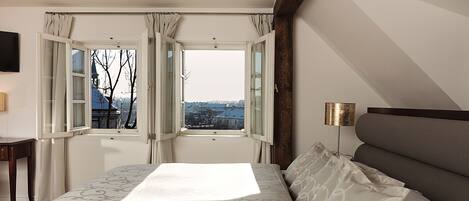 A bedroom with a large bed, a nightstand, a lamp, and a view of the city through open windows.