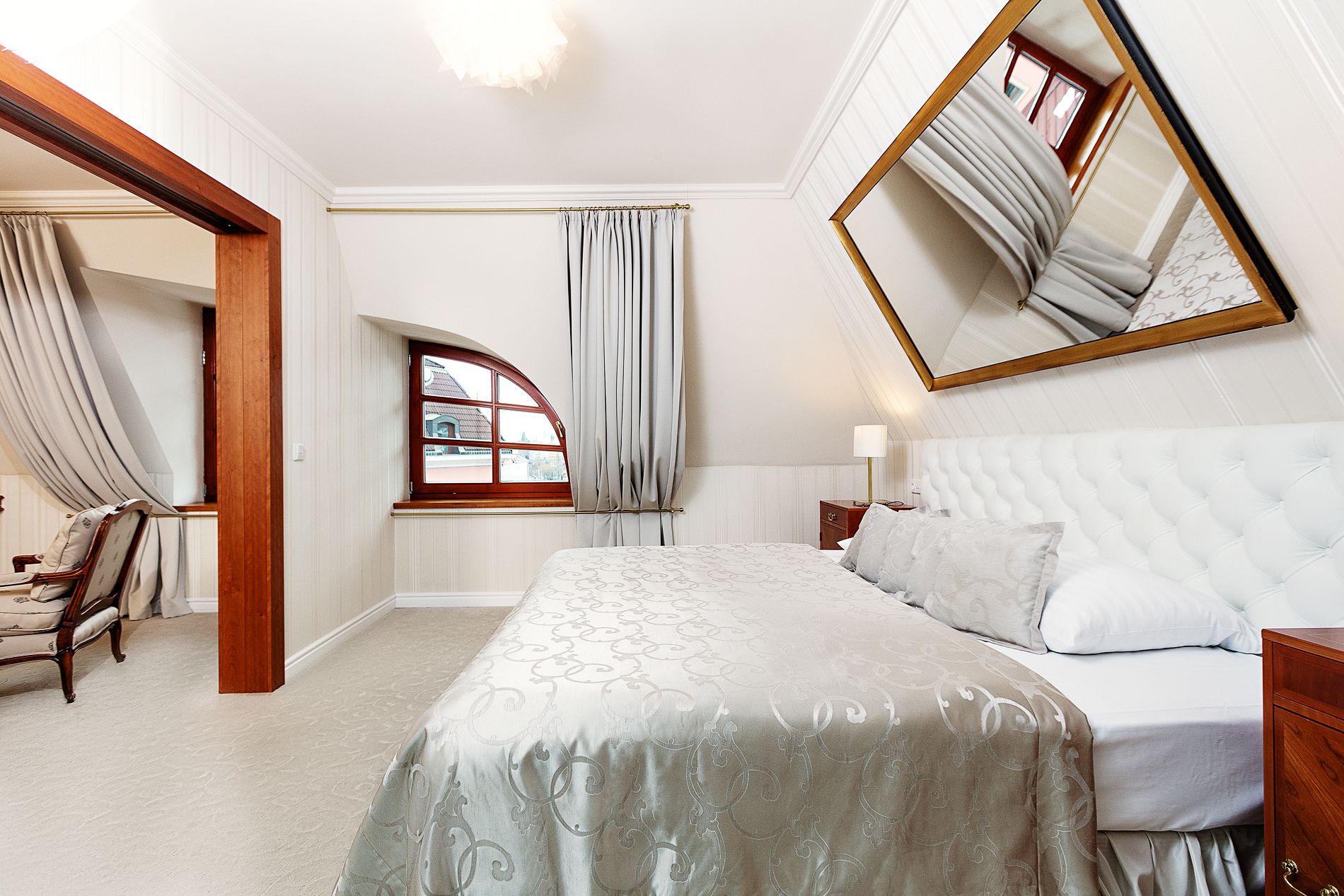 Ein Schlafzimmer mit einem großen Bett, einem Nachttisch, einem Sessel, einem Fenster mit Vorhängen und einem Spiegel an der Wand.