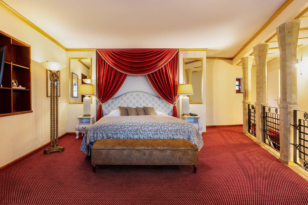 A hotel room with a large bed, red curtains, a wooden floor, a nightstand, a lamp, and a mirror.