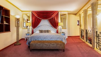 A hotel room with a large bed, red curtains, a wooden floor, a nightstand, a lamp, and a mirror.