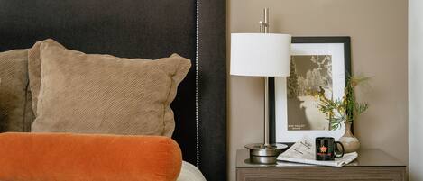 A neatly made bed with a black headboard, a brown pillow, and an orange accent pillow. Beside the bed is a small wooden nightstand with a lamp, a framed picture, a vase with a plant, and a mug.