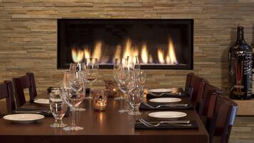 Mesa de comedor lista para seis personas, con copas de vino, platos y cubiertos, un florero decorativo y una botella de vino. Al fondo, una chimenea con fachada de piedra.