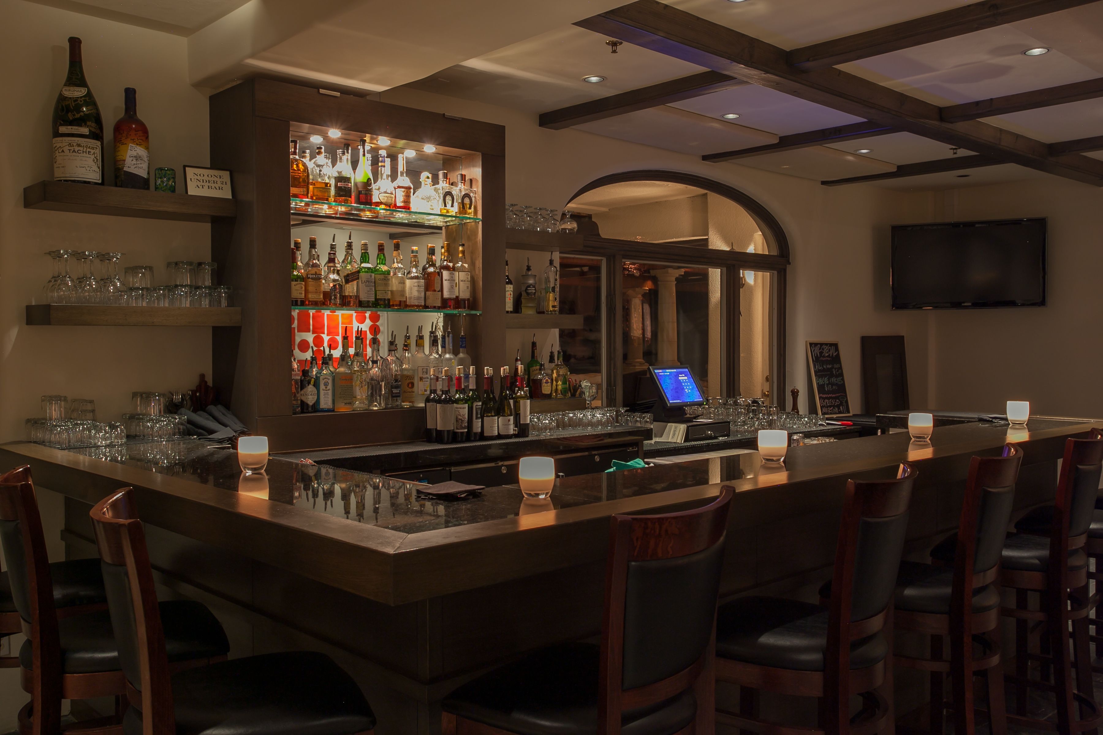A bar area with a long counter, multiple shelves with bottles, glasses, and a cash register.