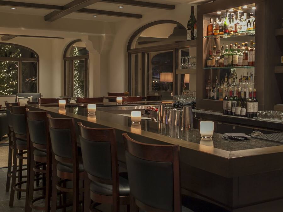 A bar area with a long counter, bar stools, and a variety of bottles and glasses on shelves.
