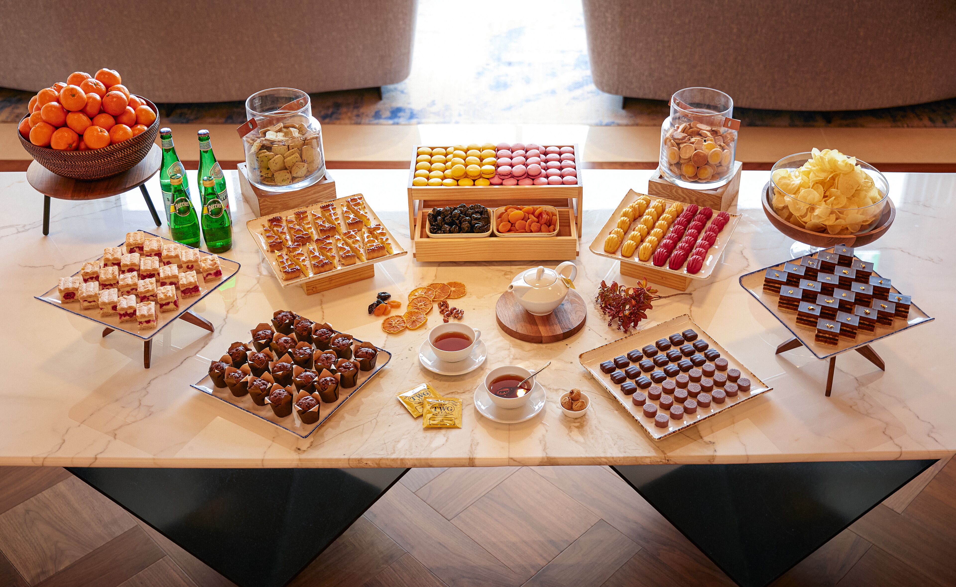 A table with various food items and drinks, including a bowl of oranges, a bottle of beer, and several trays of pastries and desserts.