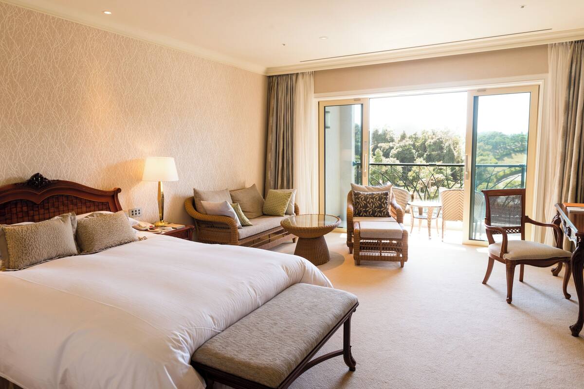 Une chambre d’hôtel spacieuse avec un grand lit, un coin salon avec un canapé et des fauteuils, et un balcon offrant une vue sur la verdure.