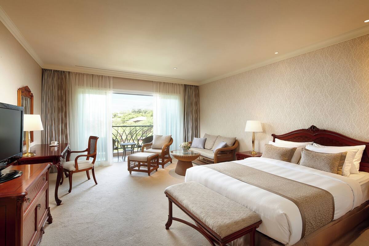Une chambre d’hôtel avec un grand lit, un bureau avec une télévision, un canapé et un fauteuil. Il y a un balcon avec des rideaux.