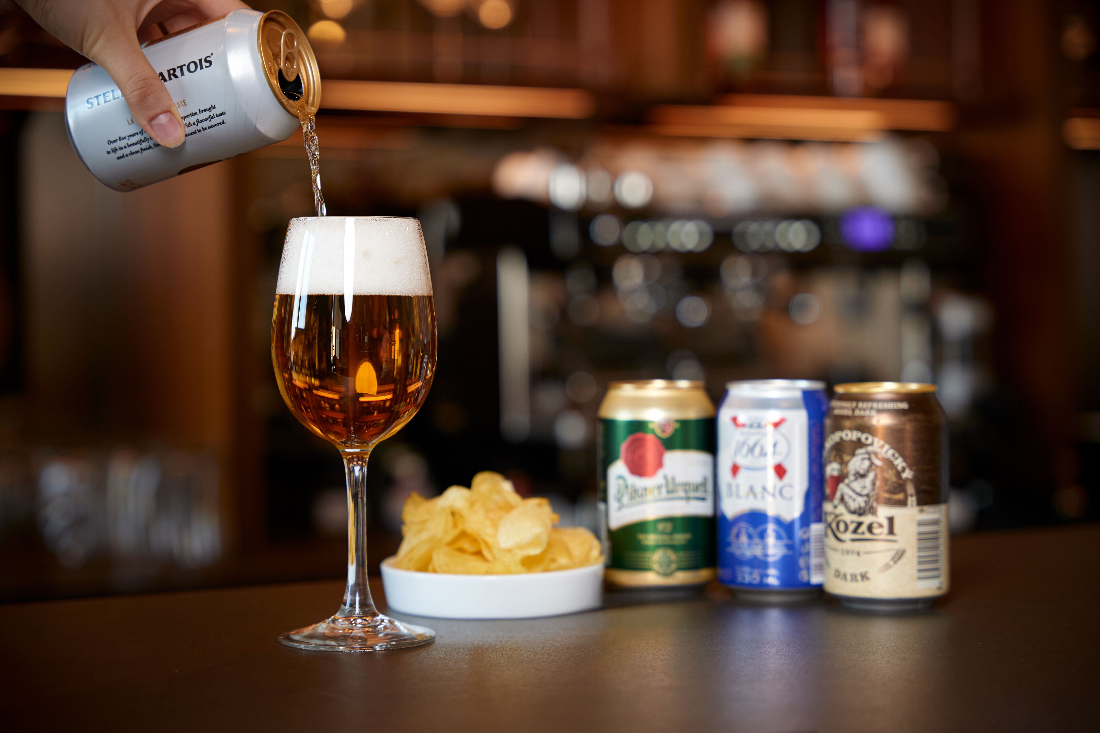 Ein Bierglas wird gerade mit Bier gefüllt, dazu liegen Chips und drei Bierdosen auf dem Tisch.