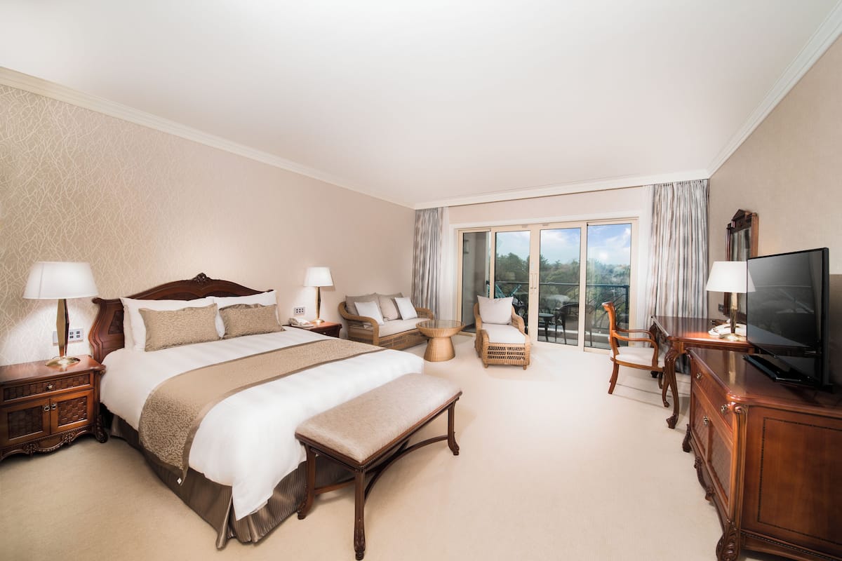Une chambre d’hôtel spacieuse, dotée d’un grand lit, d’un coin salon avec un canapé et des fauteuils, d’une télévision et d’un balcon offrant une vue agréable.