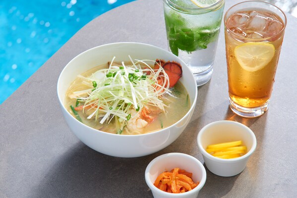A bowl of soup with vegetables and noodles, a glass of iced tea with a lemon slice, and two small bowls of pickled vegetables. - Lotte Hotel Jeju (Seogwipo)