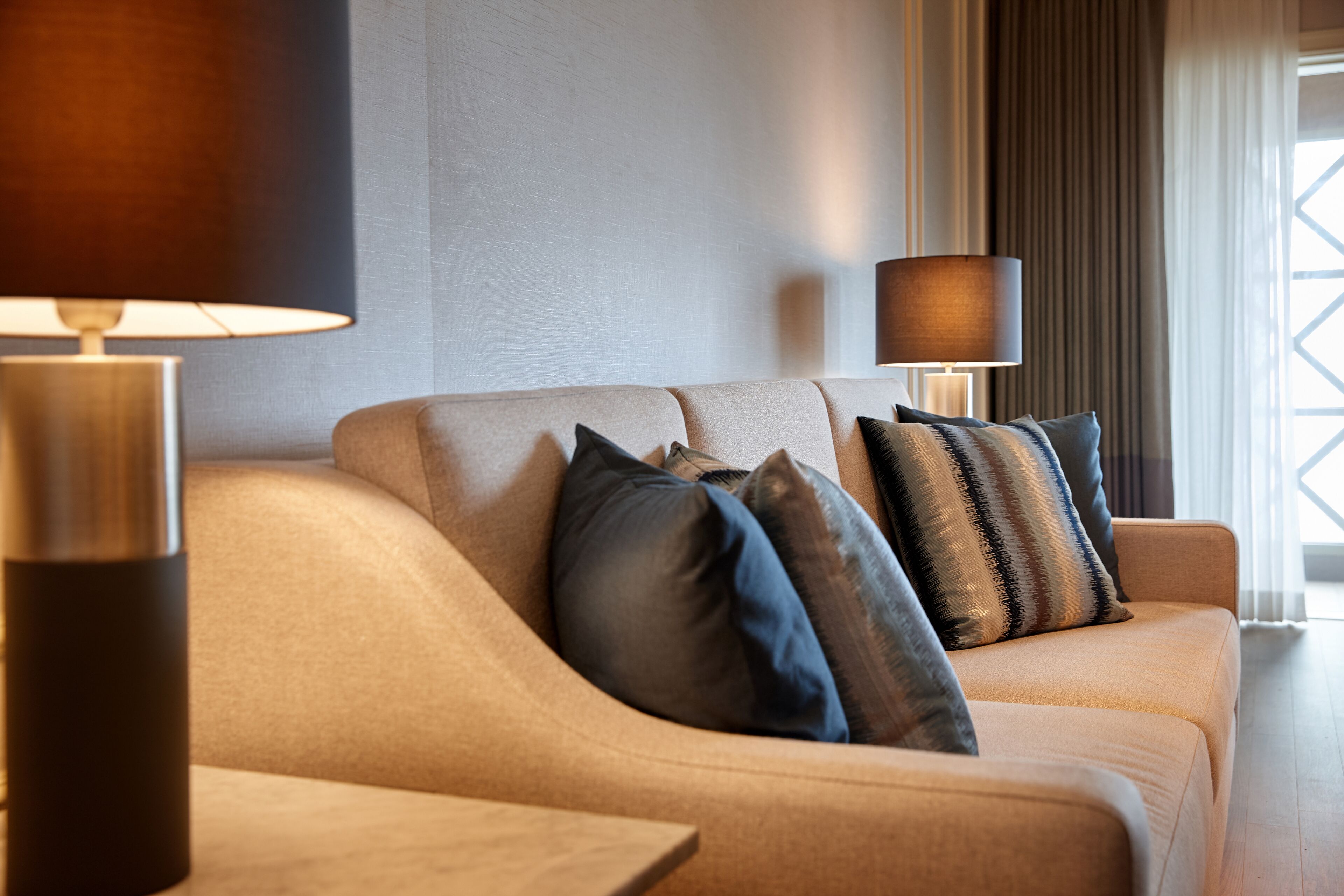 A modern living room with a beige sofa, two lamps, and a window with curtains.