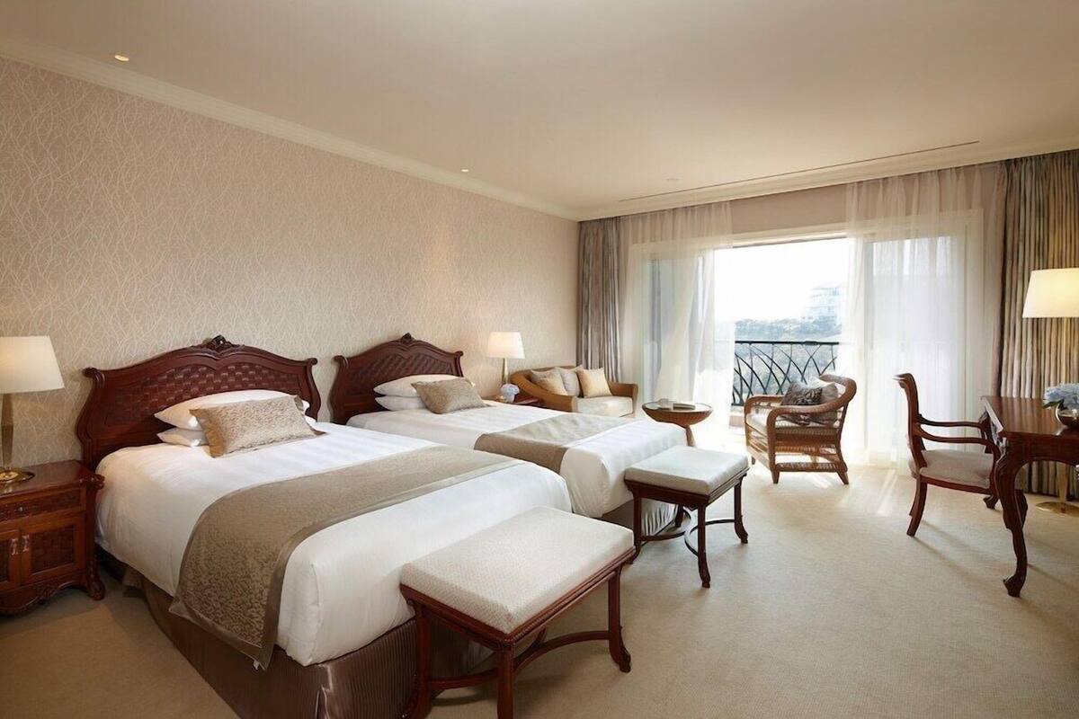 Une chambre d’hôtel avec deux lits, un coin salon comprenant une chaise et une table, et un balcon avec une balustrade.
