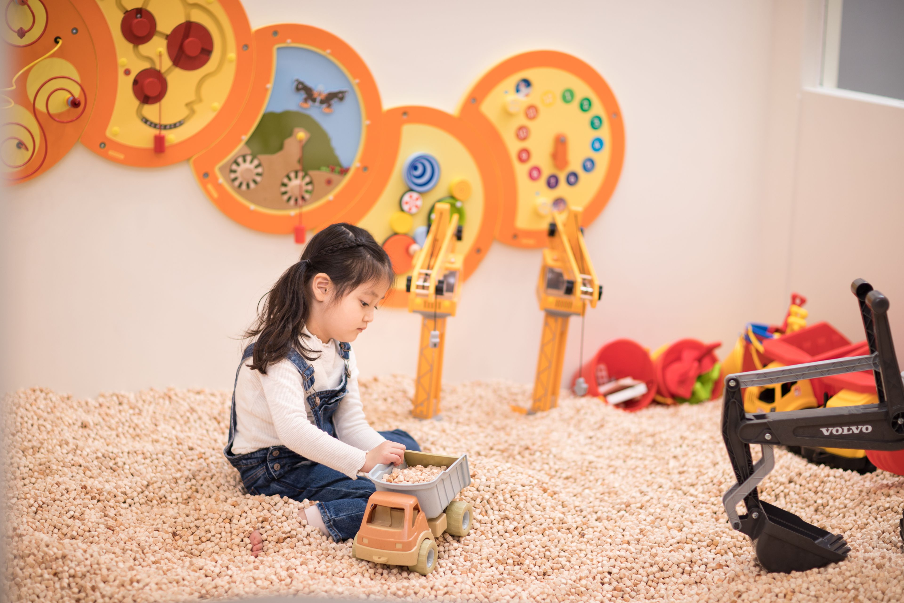 Un enfant joue avec des véhicules miniatures et une pelleteuse jouet dans une pièce dont les murs sont décorés d’éléments à caractère éducatif.