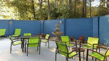 Een terras buiten met groene stoelen en tafels, een stenen fontein en een blauwe muur.