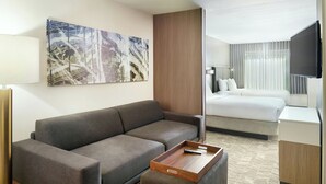 A modern hotel room with a gray sofa, a wooden side table, a bed with white bedding, a flat-screen TV mounted on the wall, and a large abstract painting above the sofa. - Springhill Suites By Marriott Atlanta Six Flags (Lithia Springs)