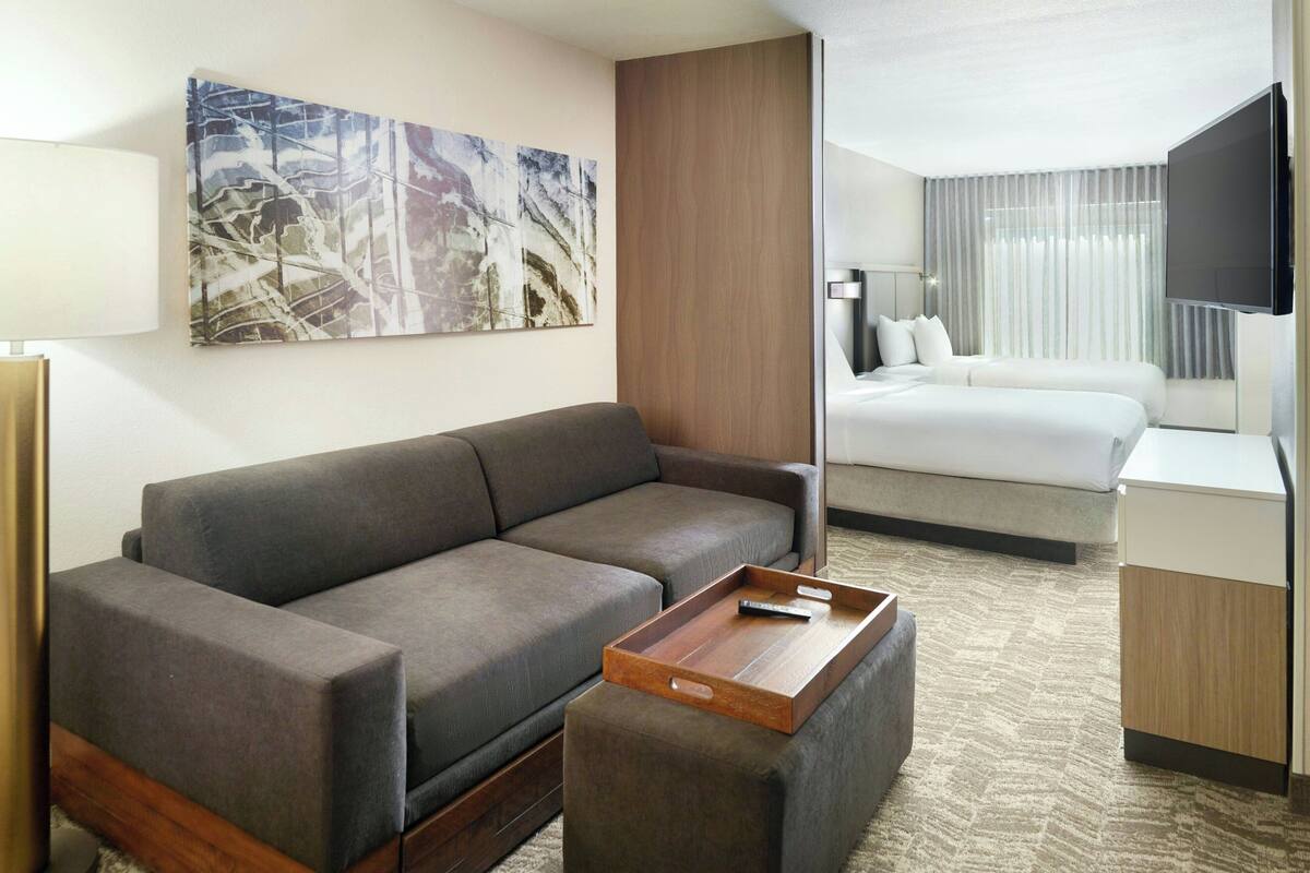 Ett modernt hotellrum med en grå soffa, ett bord i trä, en säng med vita sängkläder, en platt-TV som är monterad på väggen och en stor, abstrakt tavla ovanför soffan.