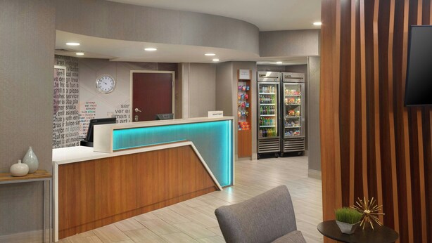 A hotel reception area with a wooden reception desk, a clock, a refrigerator stocked with beverages, and a television on the wall. - Springhill Suites By Marriott Atlanta Six Flags (Lithia Springs)