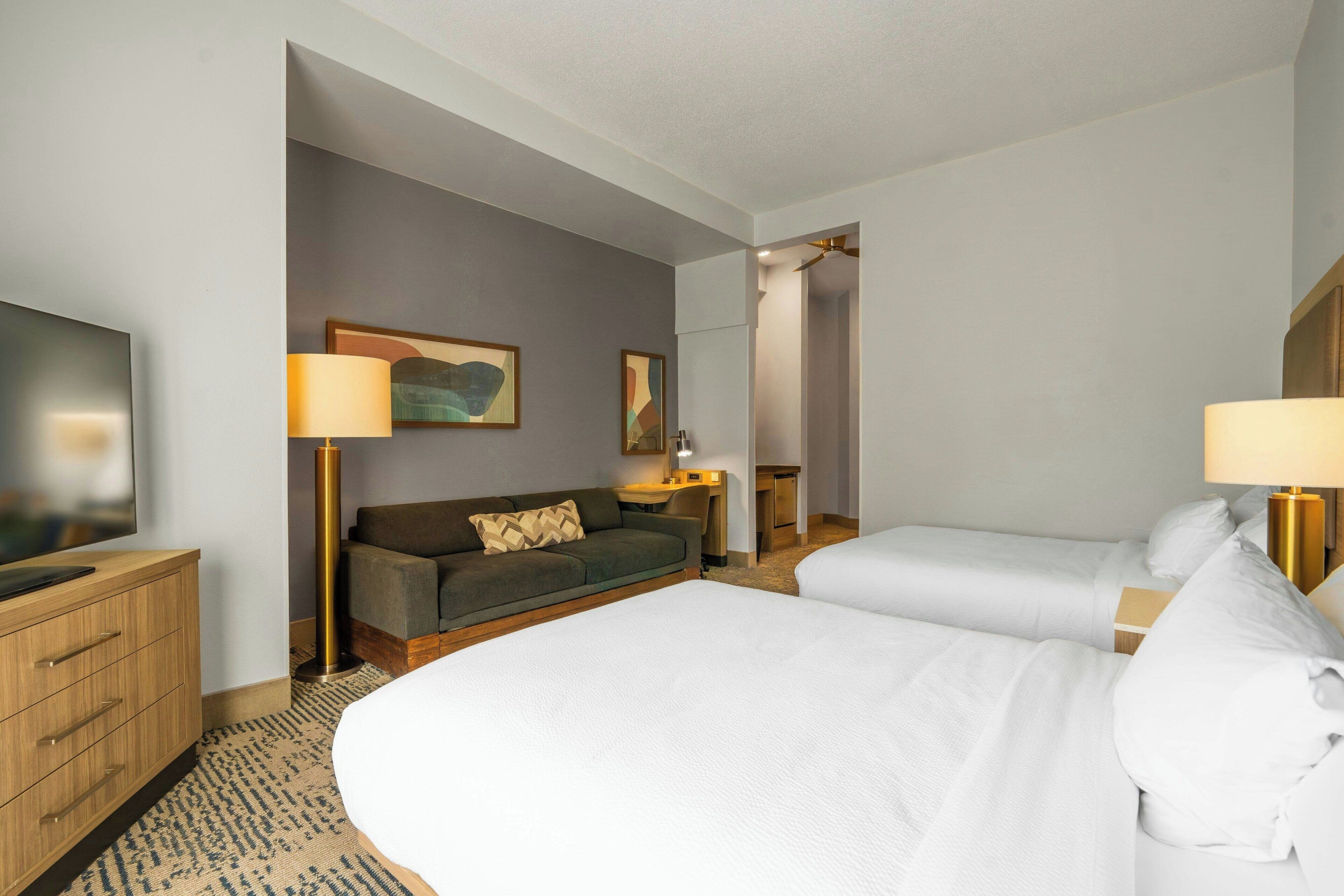 A hotel room with two beds, a sofa, a desk, a TV, and a painting on the wall.