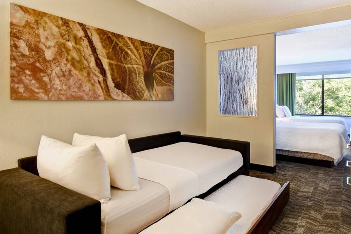 A hotel room with a sofa bed, a large abstract painting, and a view of another room with a bed.