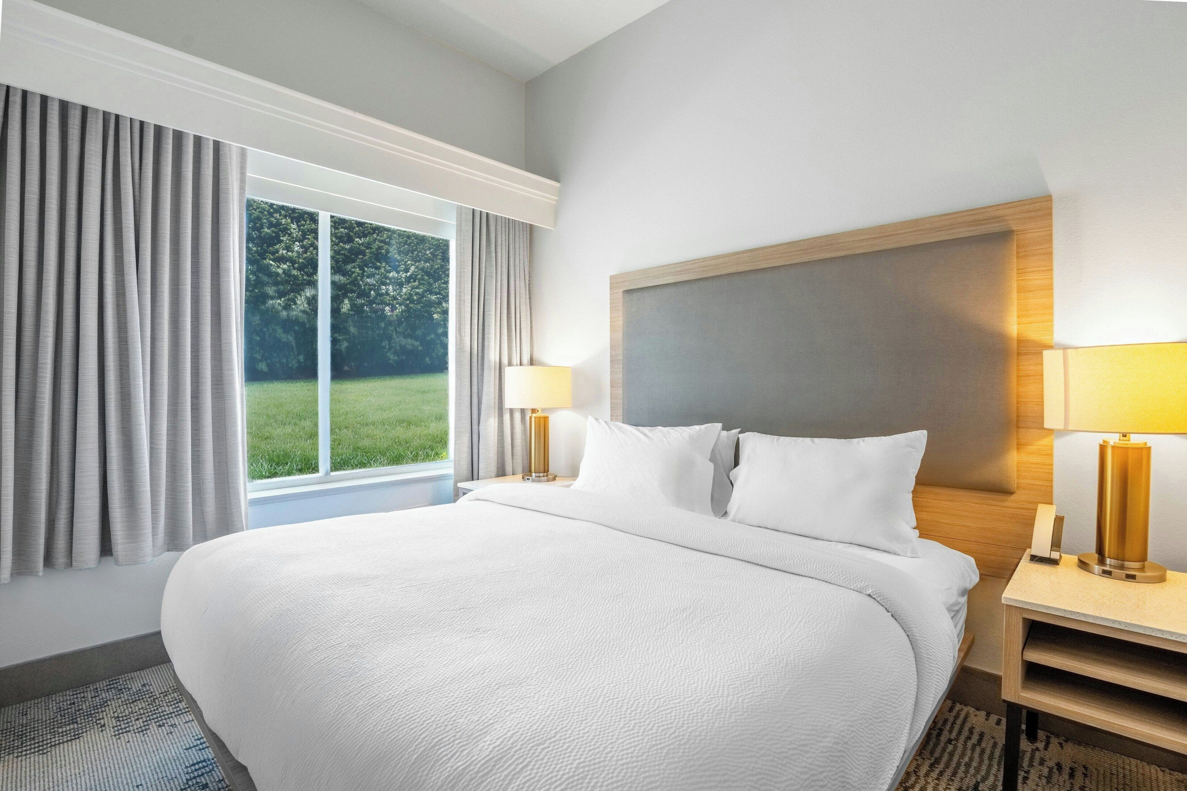 A large bed with white bedding, two bedside lamps, a nightstand, and a window with curtains.