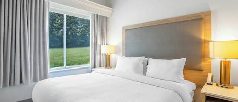 A large bed with white bedding, two bedside lamps, a nightstand, and a window with curtains.