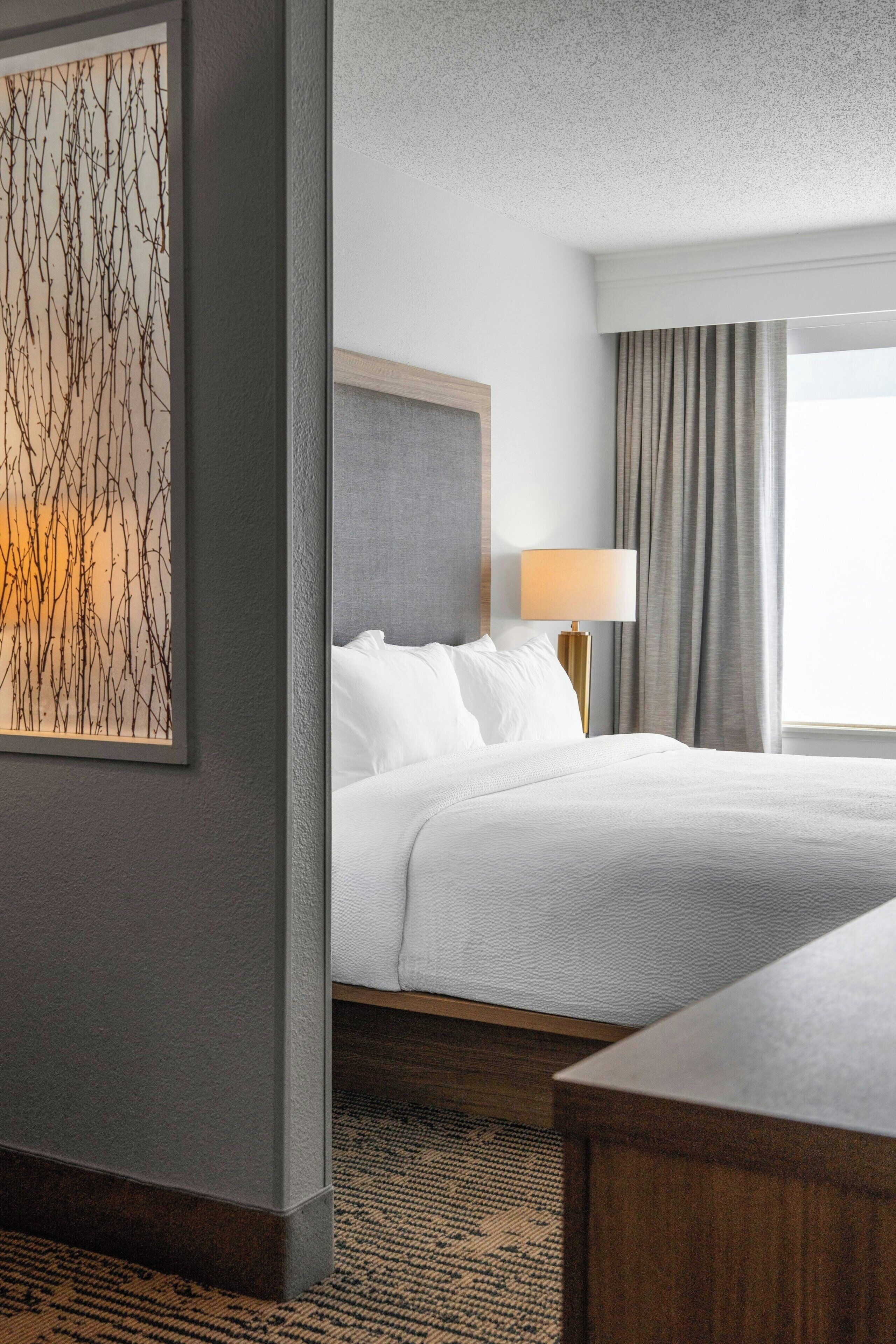 Habitación de hotel con cama, mesita de noche, lámpara, ventana con cortinas y una obra de arte en la pared.