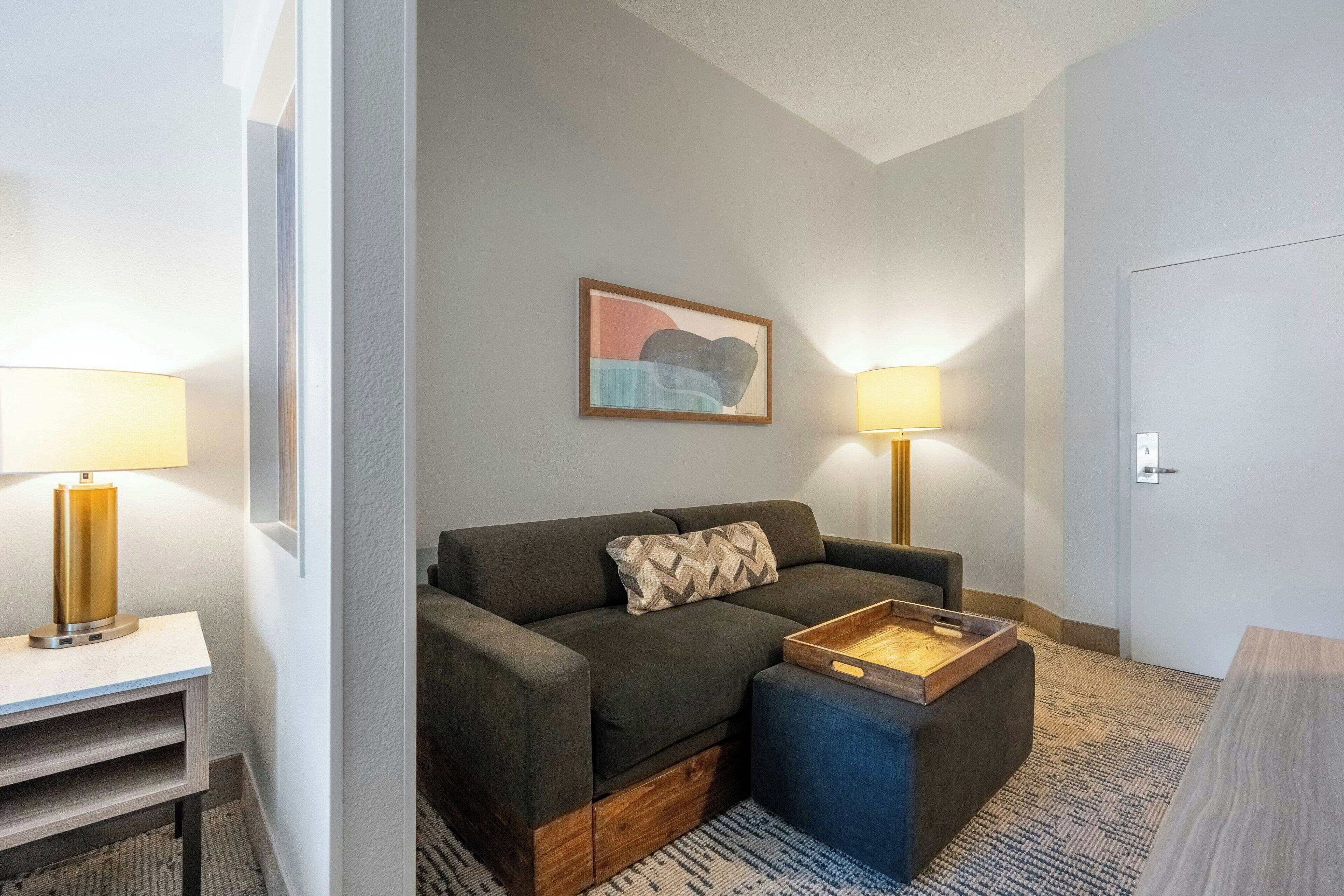 A modern hotel room with a sofa, ottoman, and a small wooden tray table. There are two wall-mounted lamps and a framed abstract painting on the wall.