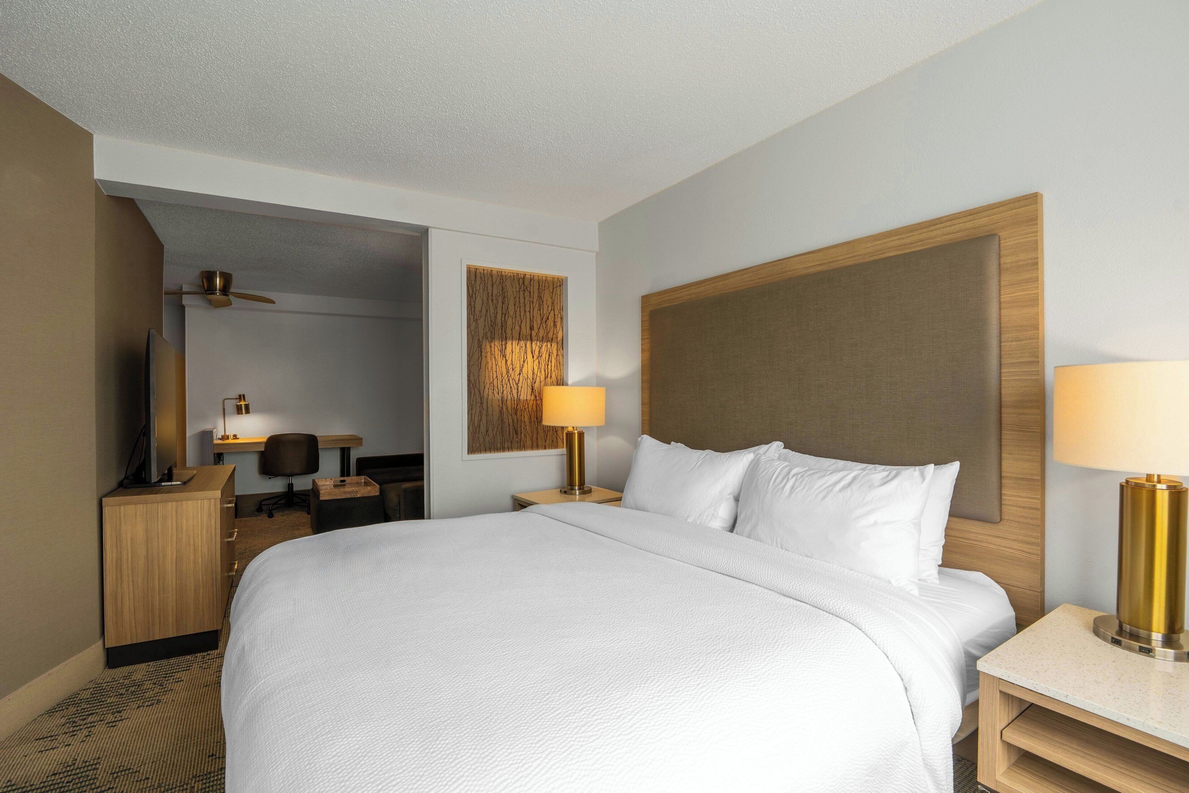 Habitación de hotel con una cama grande, un escritorio, una silla, una mesita de noche con lámpara y un ventilador de techo.