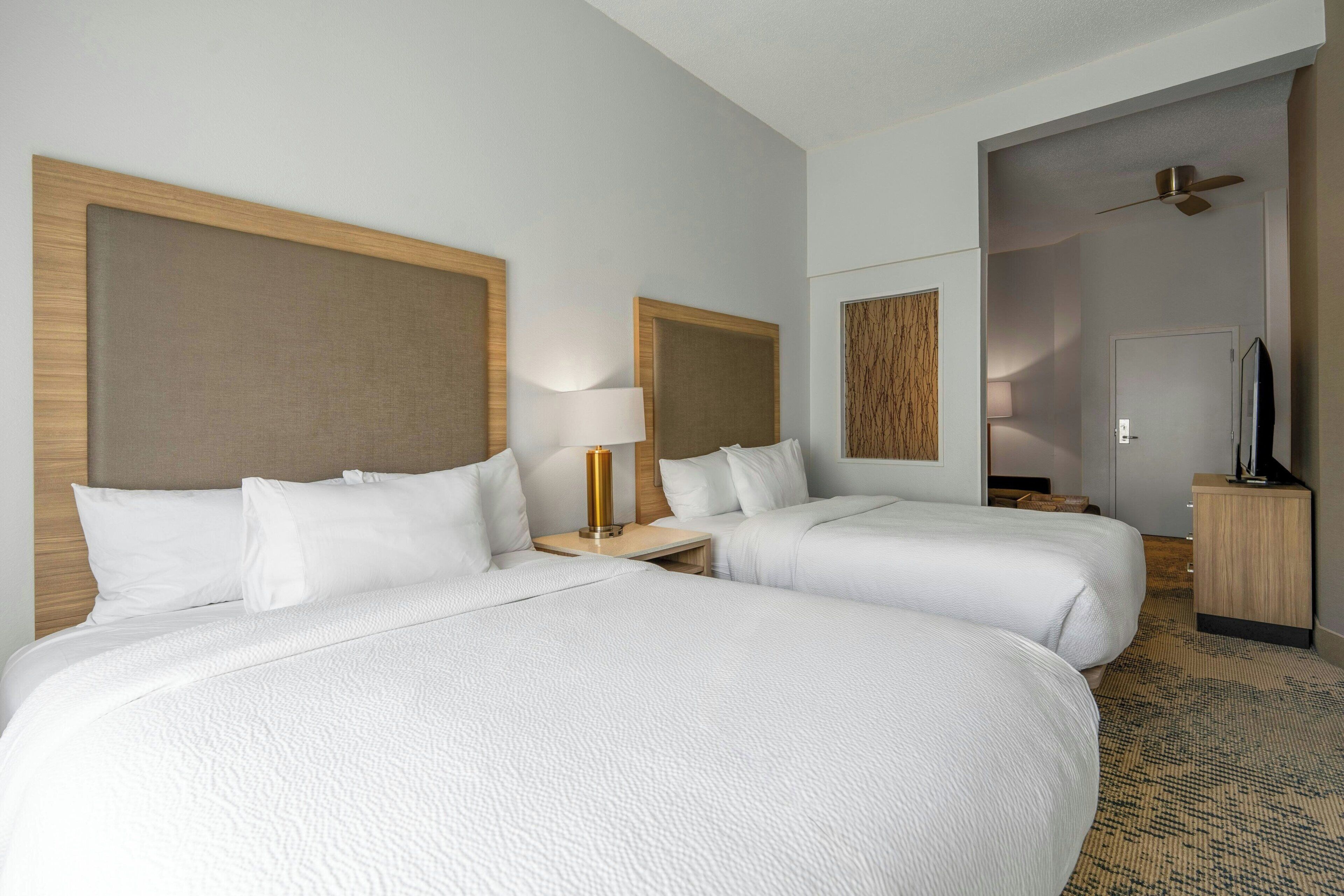 Une chambre d’hôtel avec deux lits, une télévision, un ventilateur de plafond et une tête de lit en bois.
