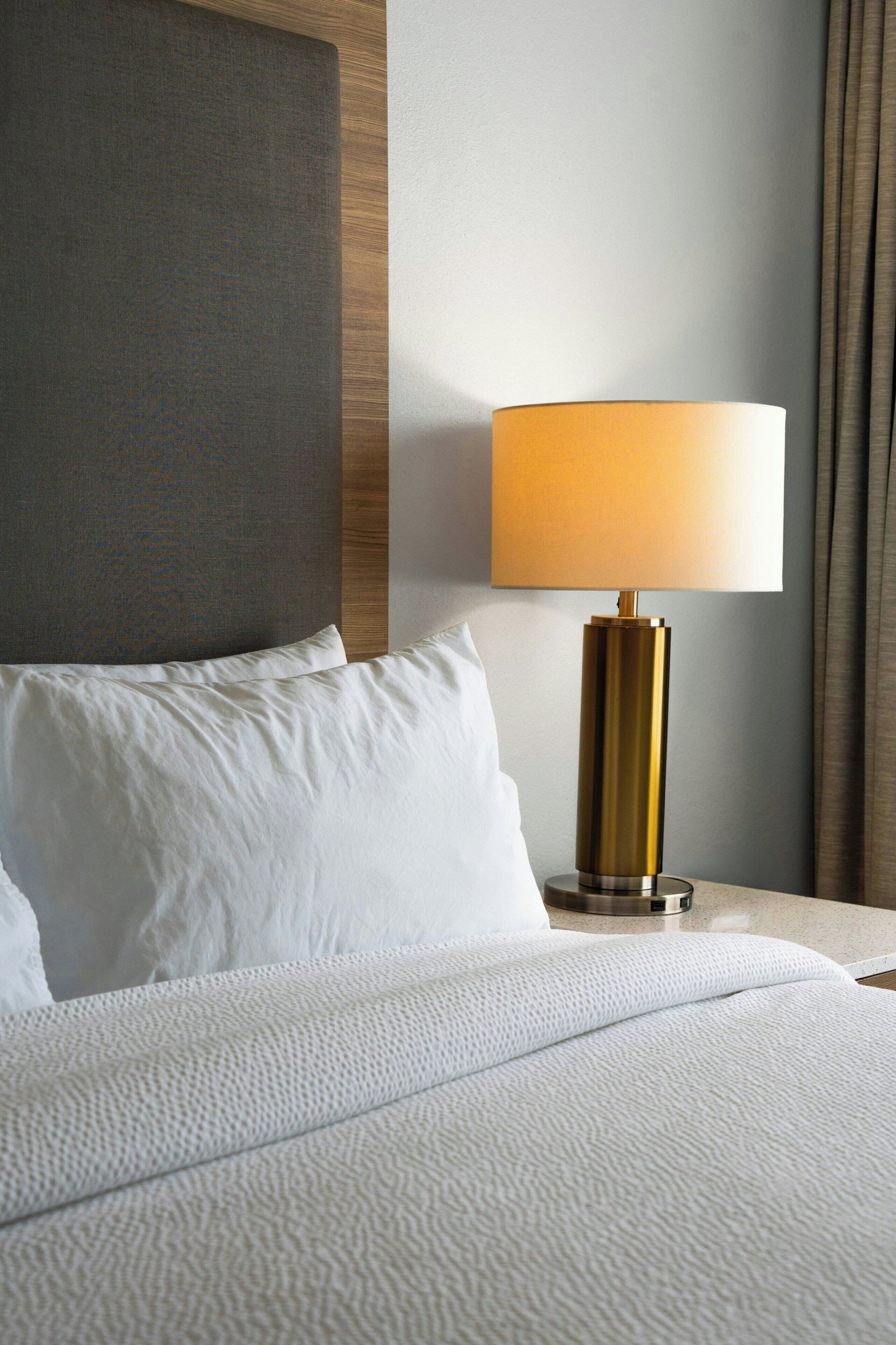 A neatly made bed with white linens, a gold-colored lamp on a bedside table, and a wooden headboard.