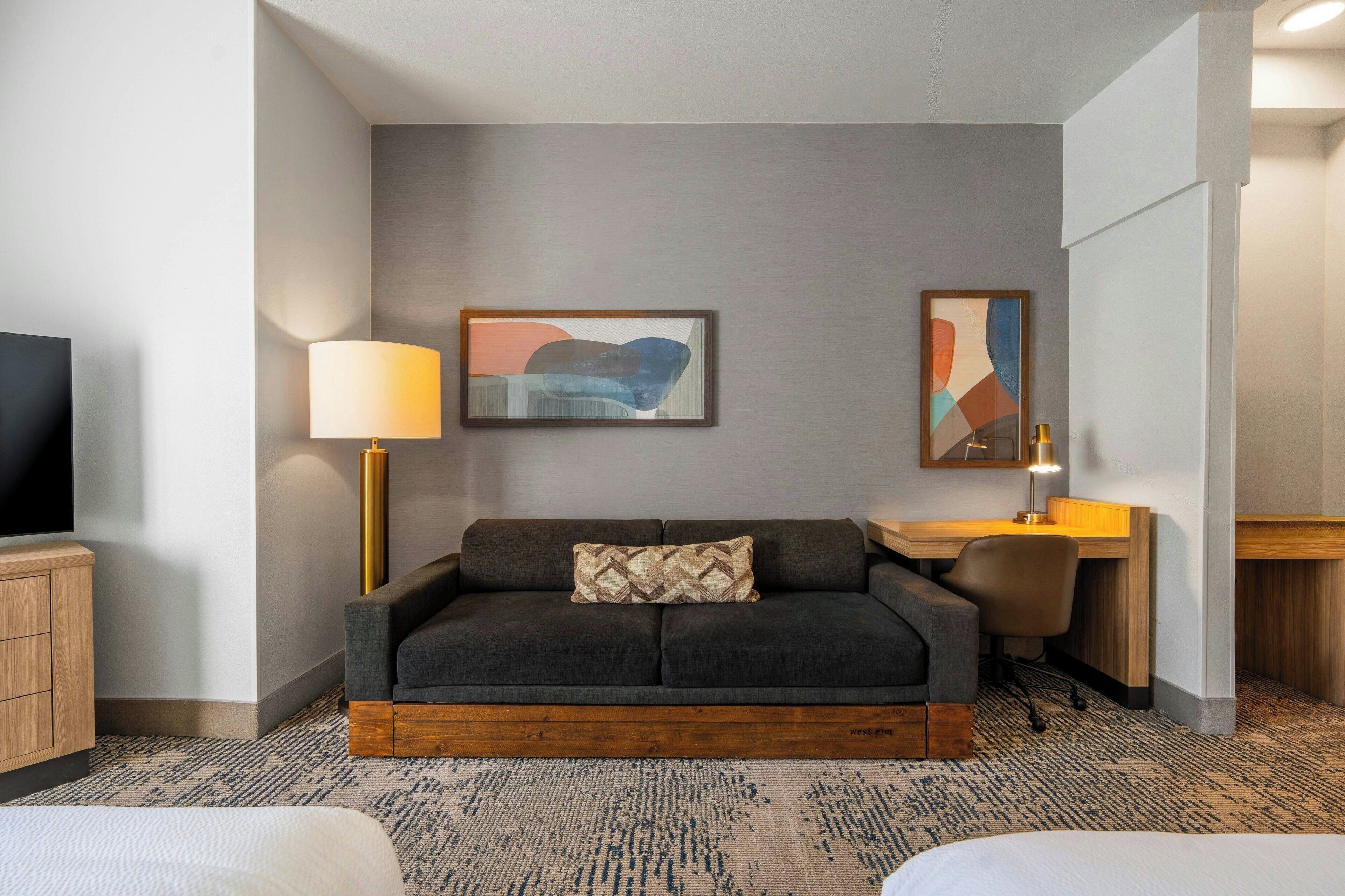 A hotel room with a sofa, a desk, a lamp, and a TV.