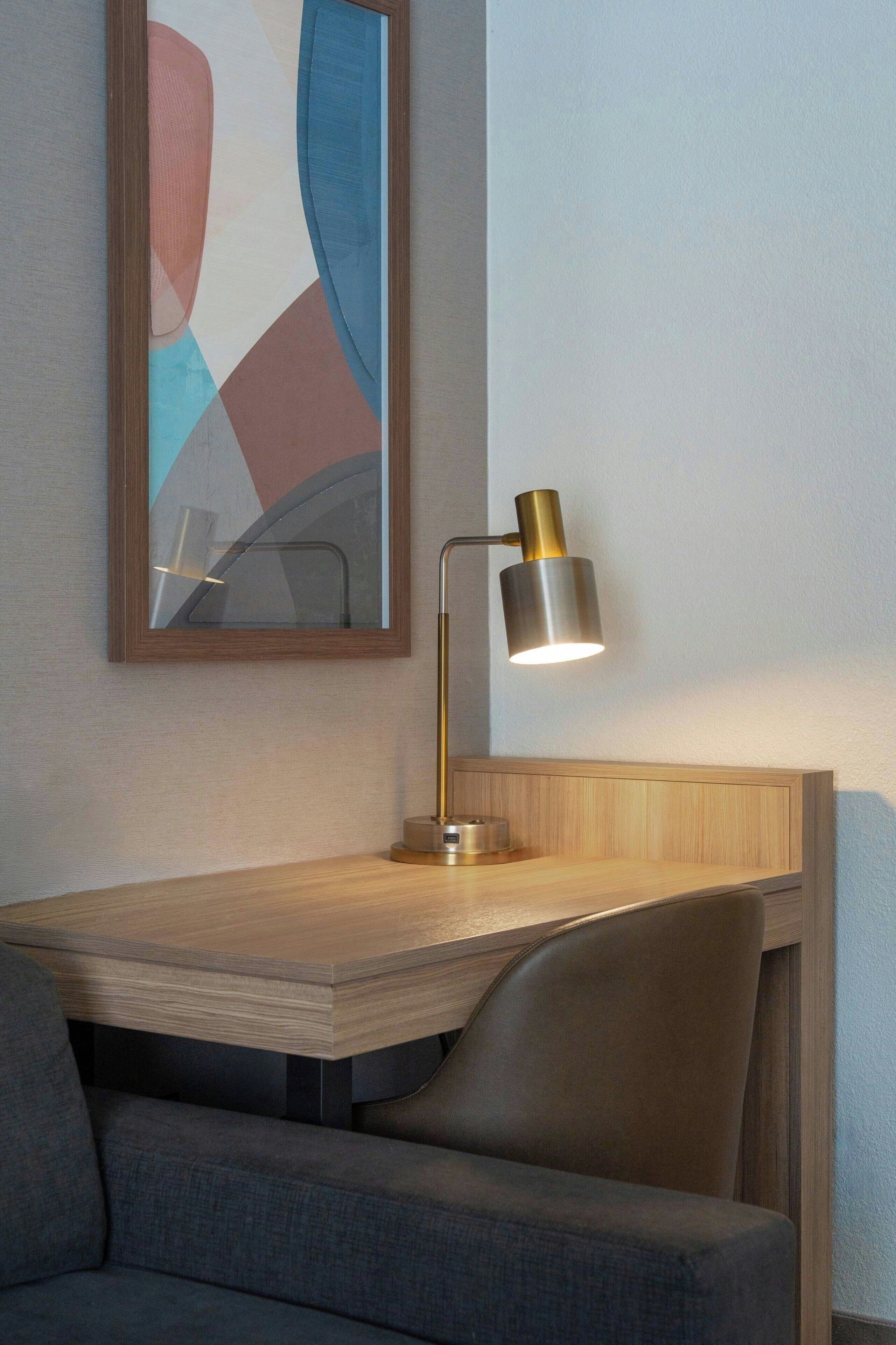 Una habitación moderna con un escritorio de madera, una silla, una lámpara y un cuadro abstracto enmarcado en la pared.