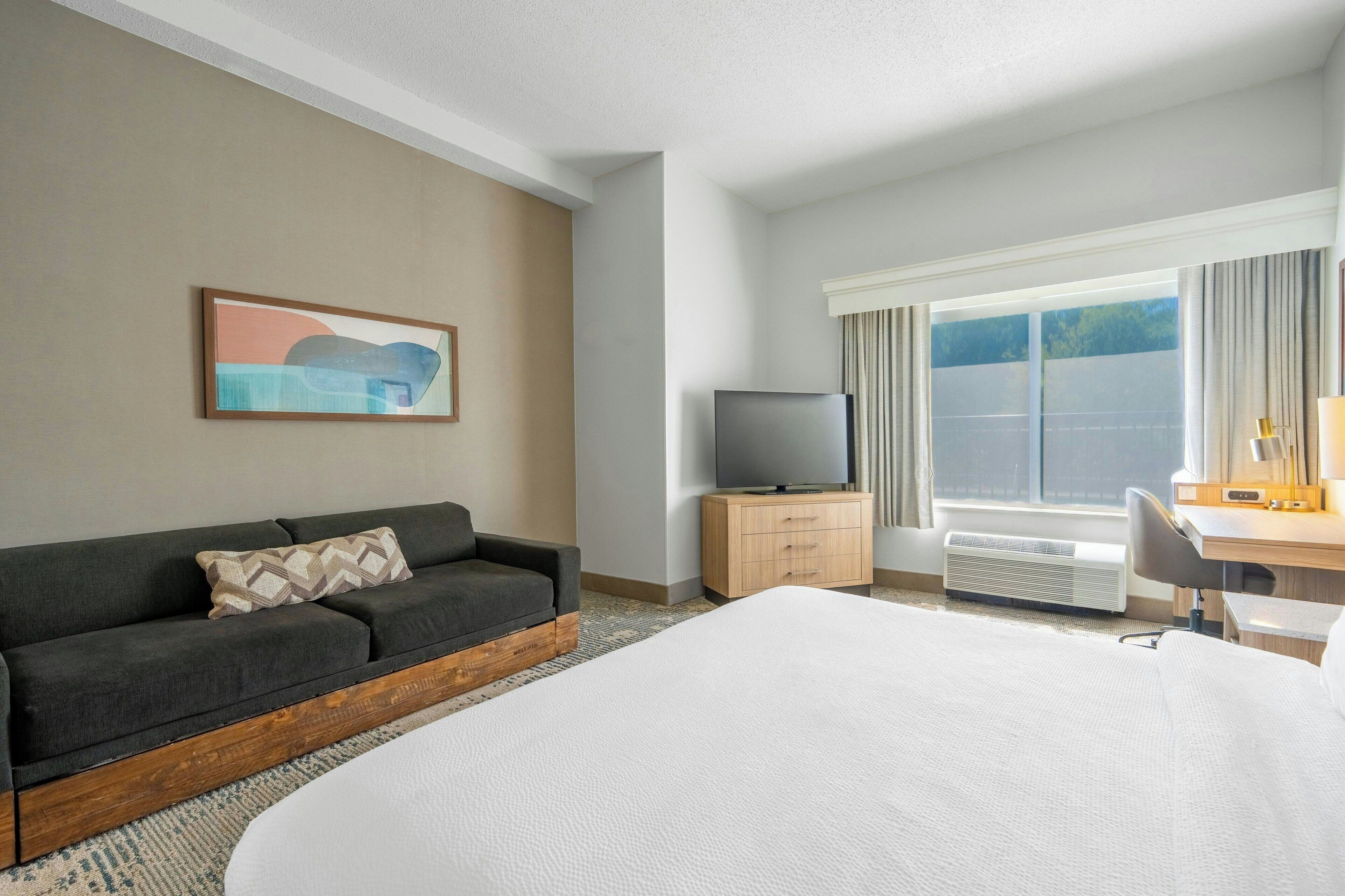 Ein Hotelzimmer mit Bett, Sofa, Fernseher, Schreibtisch und einem Fenster mit Vorhängen.