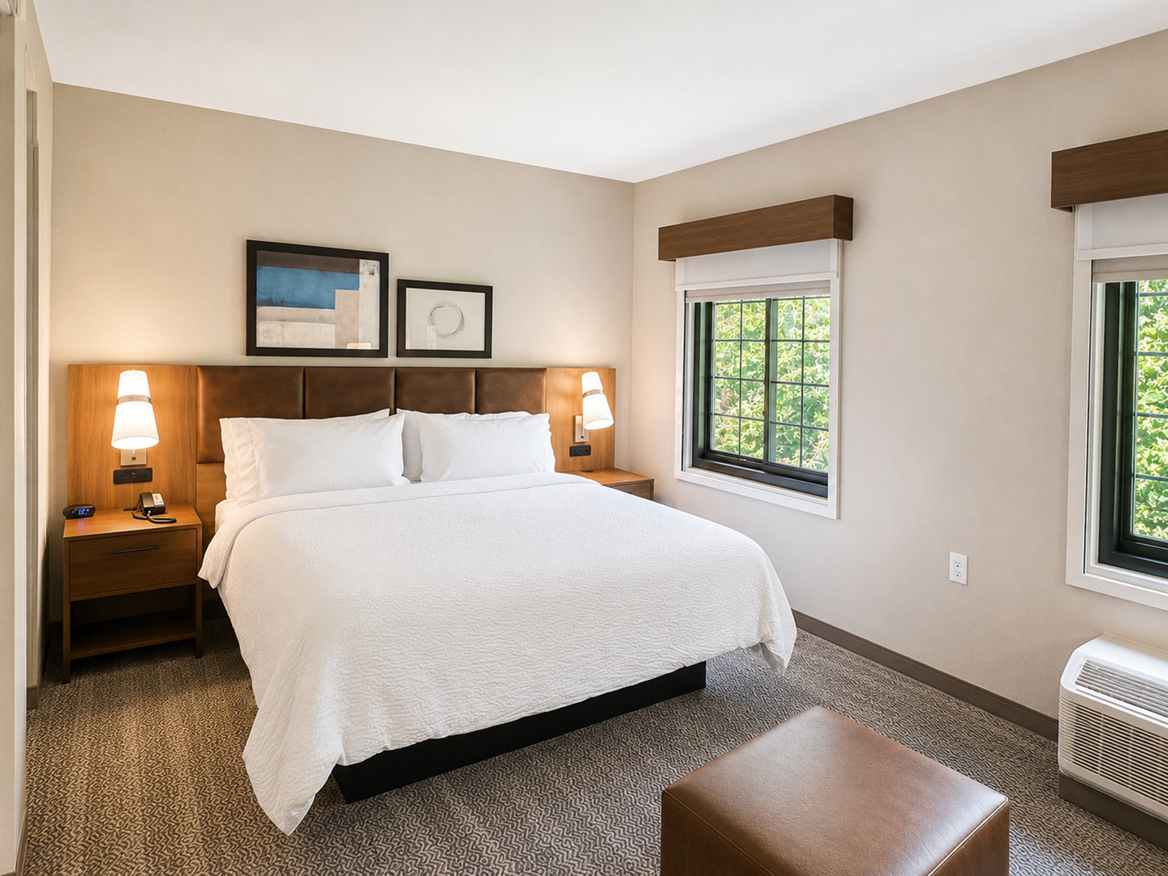 A hotel room with a large bed, two bedside tables with lamps, a window with a view of greenery, and a wall-mounted air conditioner.