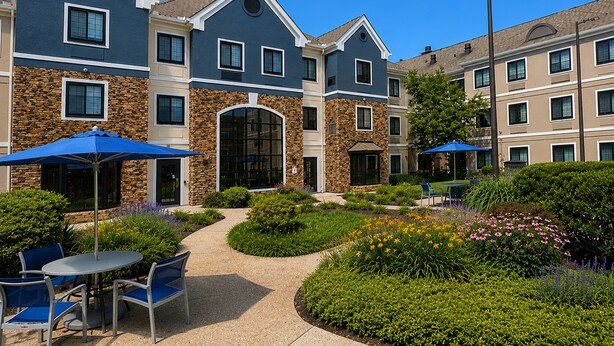 A multi-story hotel with a stone facade, outdoor seating area with blue umbrellas, and a landscaped garden. - Staybridge Suites Columbia Baltimore by IHG (Columbia)