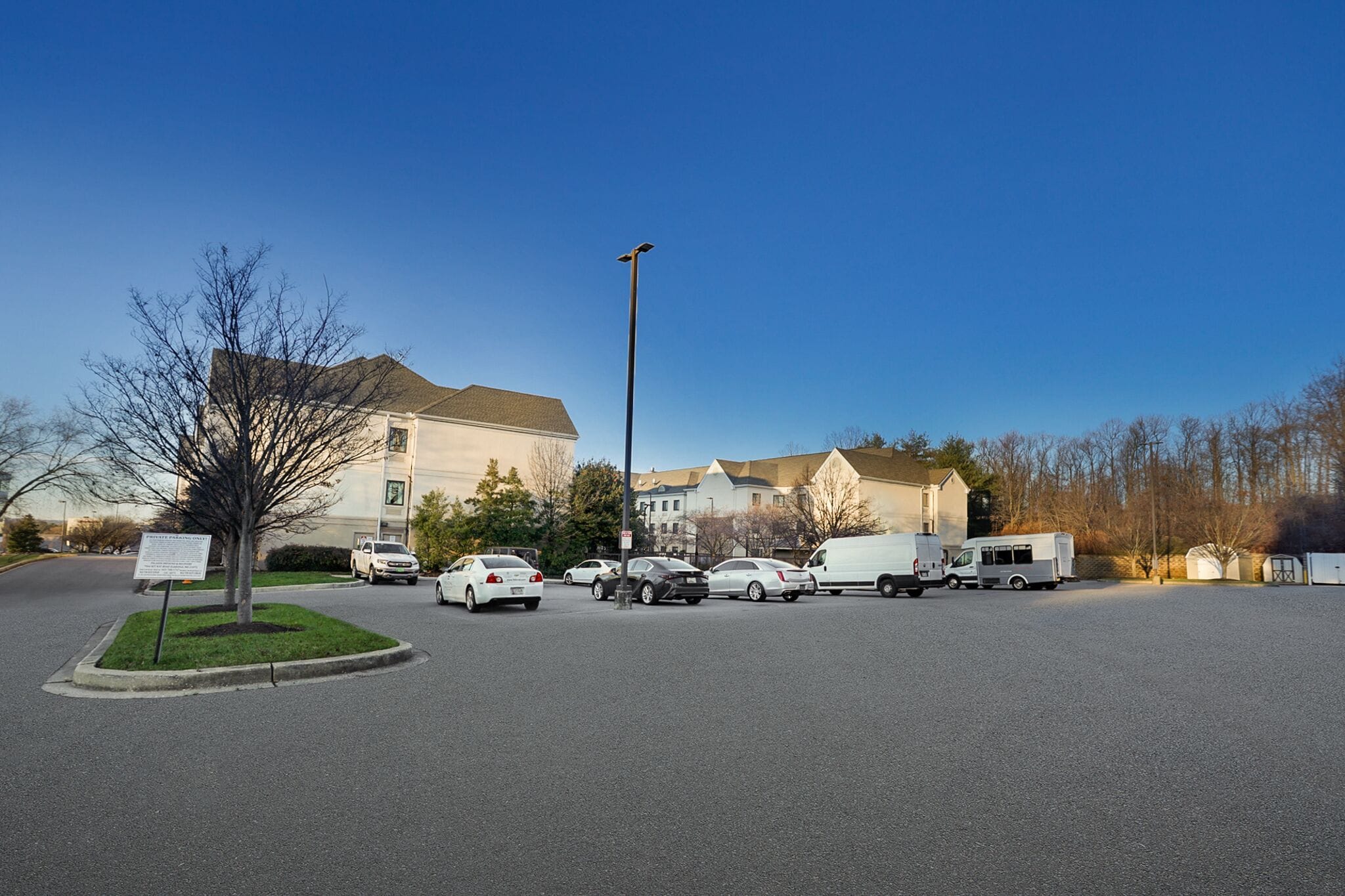 A parking lot with several cars and a white van, surrounded by buildings and a clear sky.