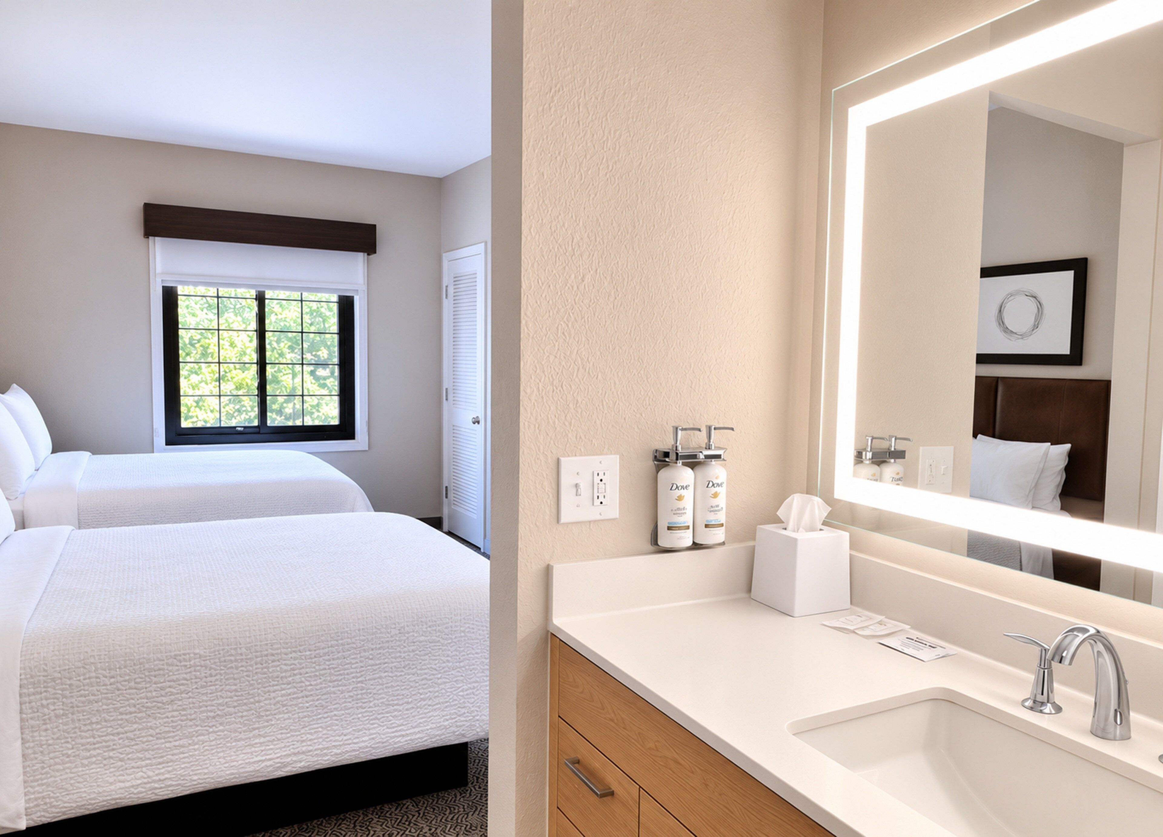 A hotel room with two beds, a window, a bathroom with a large mirror, and toiletries on the counter.