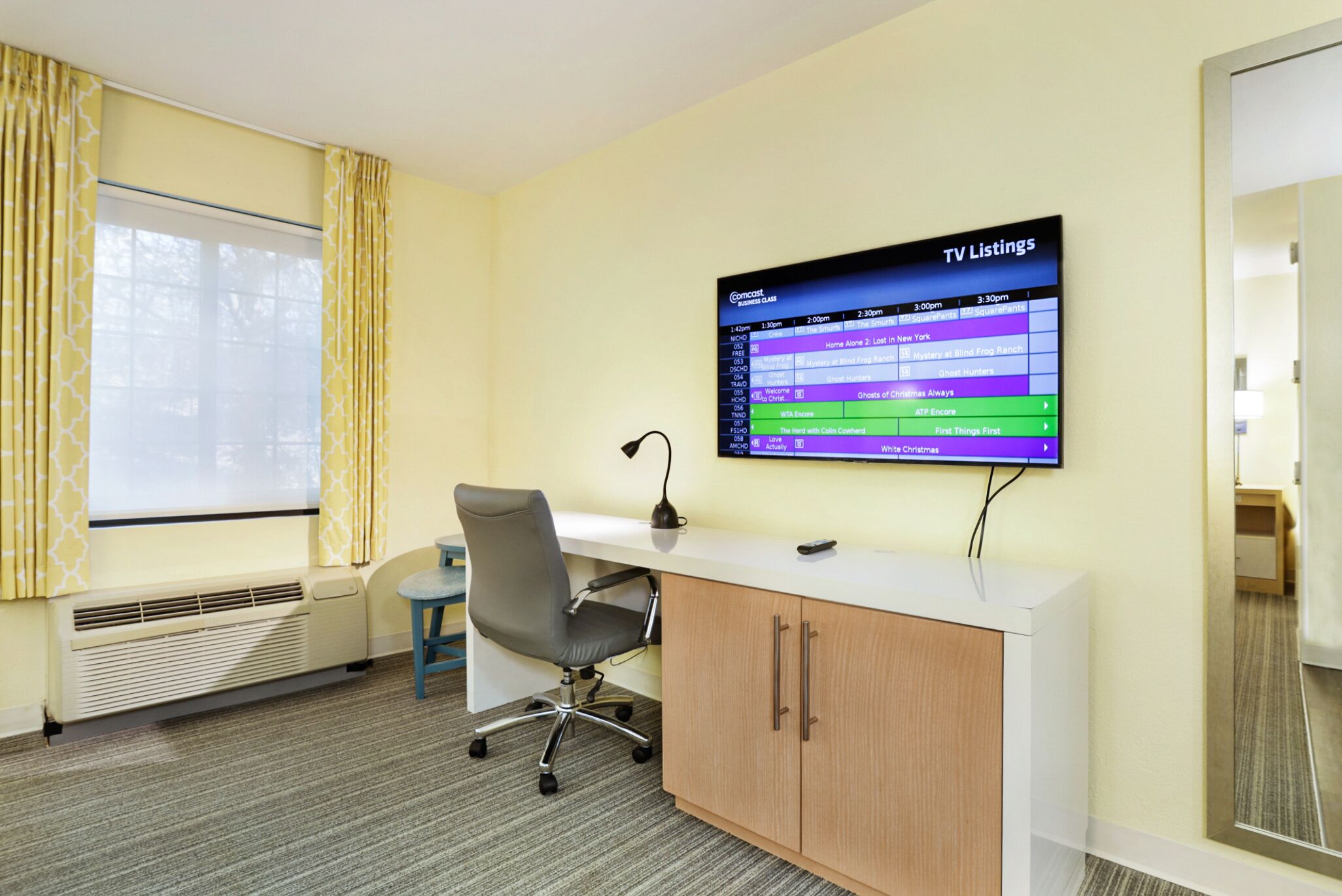 A hotel room with a TV displaying TV listings, a desk with a chair, a window with curtains, and a radiator.