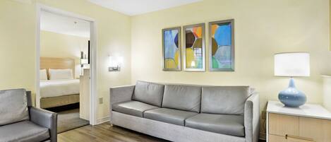 A hotel room with a grey sofa, a small armchair, a bedside table with a lamp, and a bed with white bedding. There are abstract paintings on the wall and a wooden cabinet.