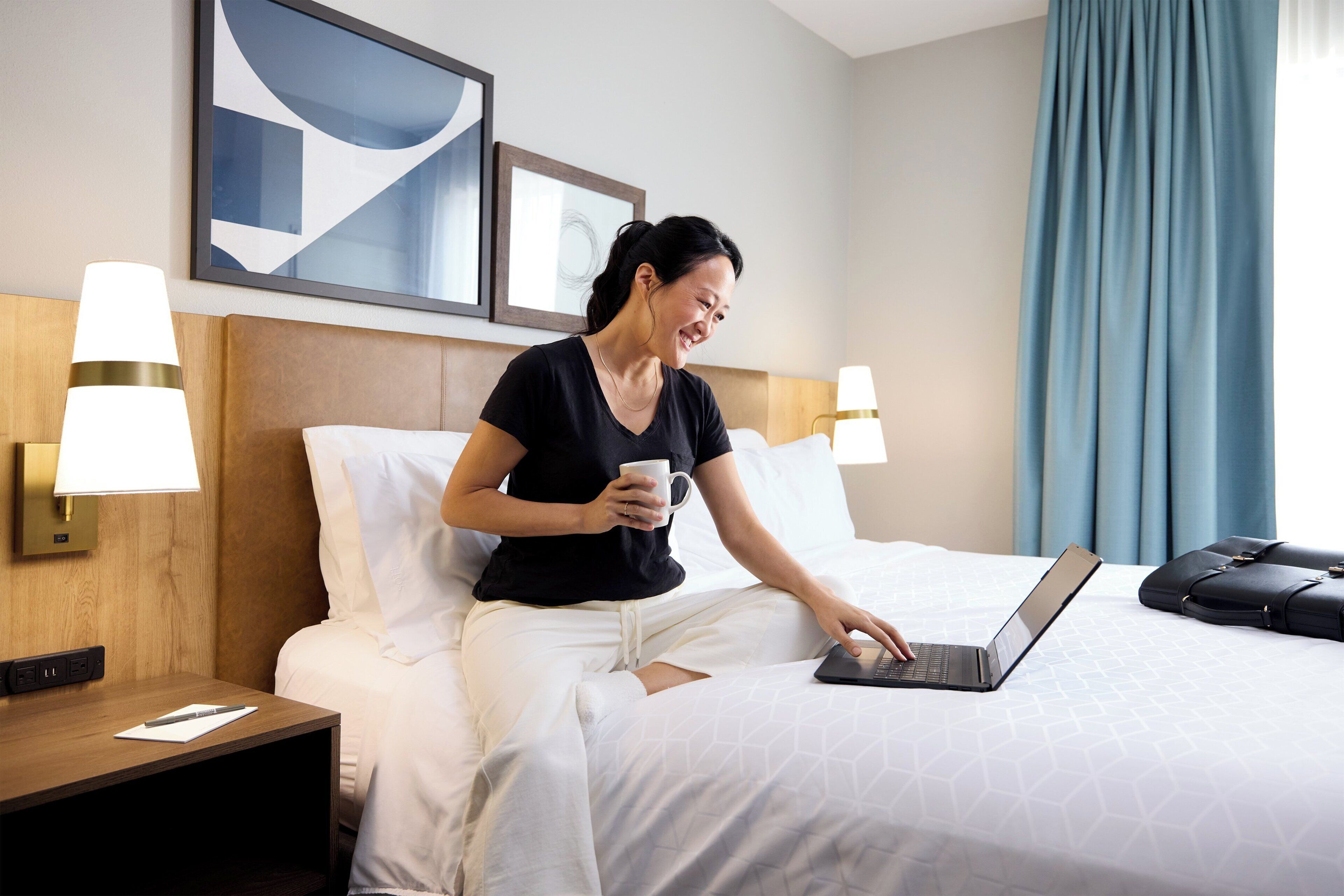 A hotel room with a bed, a laptop, a mug, and a suitcase.