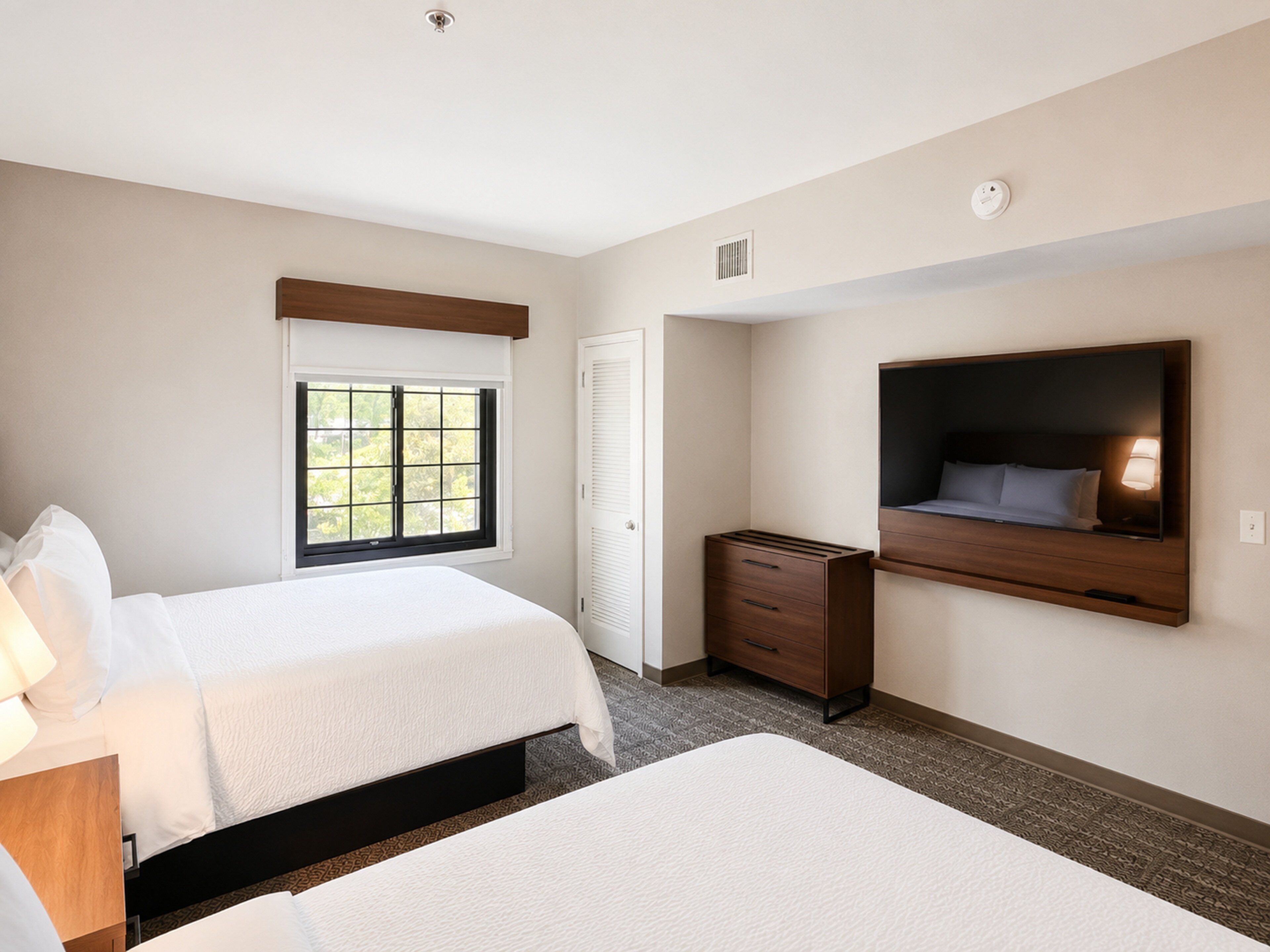A hotel room with two beds, a TV mounted on the wall, a window, and a desk.